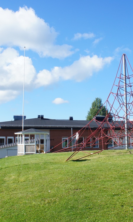 Sommarbild tagen på skolgården vid mellanstadiet med klätternät, grön gräsmatta och byggnader.