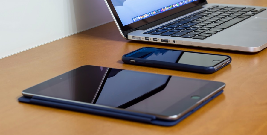 En ipad, iphone och en dator som står på ett brunt bord.