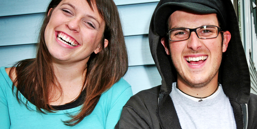 En man med glasögon och en grå tröja står bredvid en kvinna i blå tröja. Det båda står med armarna i kors framför en ljusblå vägg och skrattar tillsammans.