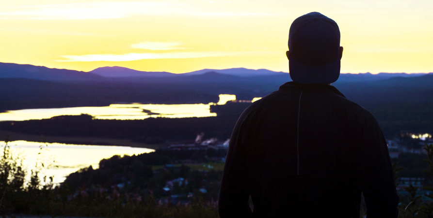 Kille med keps står på Tjamstanberget och tittar ut över Malå. Det är sommarkväll och solen har precis gått ner bakom berget.