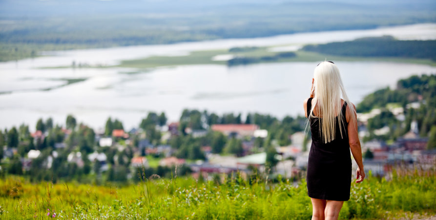 Kvinna i svart klänning med ljust långt hår blickar ut över Malås samhälle på Tjamstanbacken.