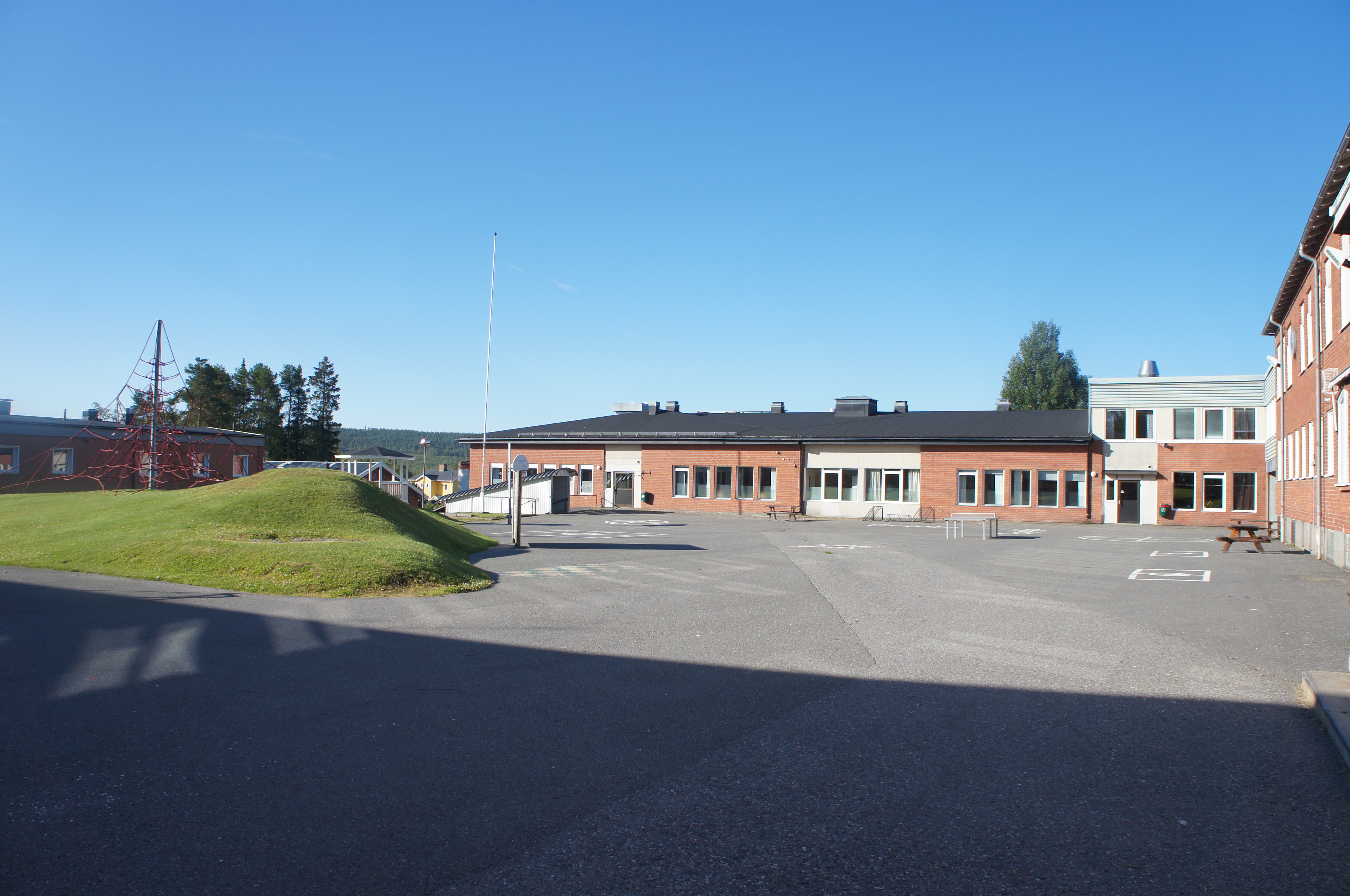 Sommarbild tagen in mot mellanstadiets skolgård med asfalterad gårdsplan, gräskulle och byggnader.