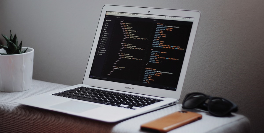 Silverfärgad bärbar dator som står öppen på ett grått underlag,  till höger ligger en mobiltelefon som ligger upp och ned, svarta solglassögon och till vänster står en blomma med vit kruka