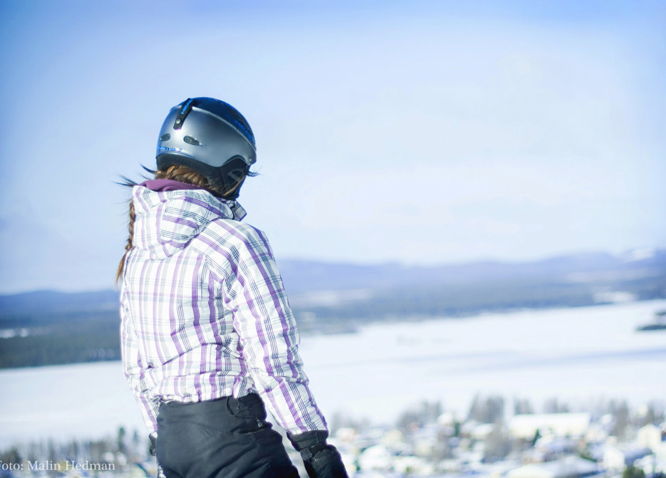 Flicka i rutig jacka och slalomhjälm som spanar ut över vintrigt Malå från toppen av Tjamstanbackarna.