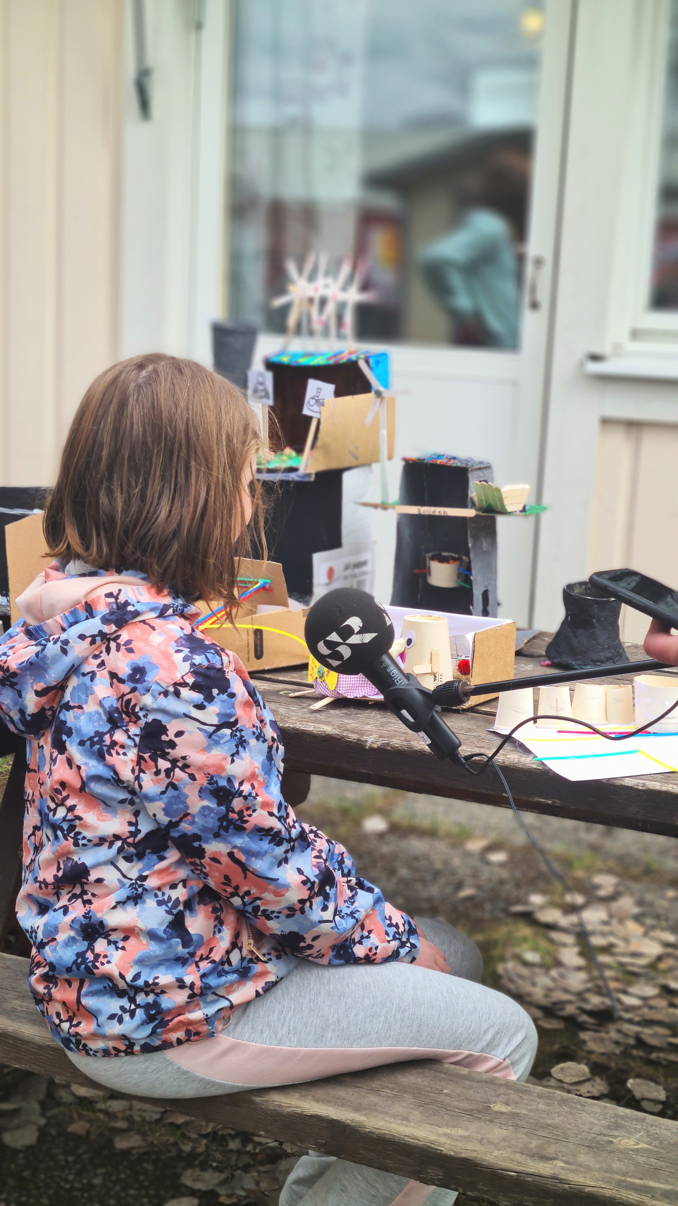 Flicka i årskurs 5 som sitter på en bänk vid ett bord utomhus och pratar i en mick med texten SR på.