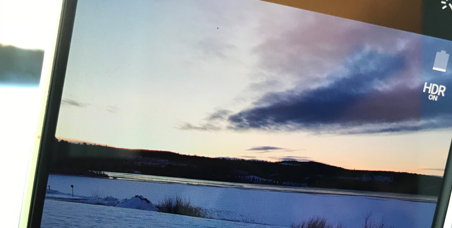 Närbild på mobiletelefon som tar ett foto på en sjö med snö och is. Berg i bakgrunden.